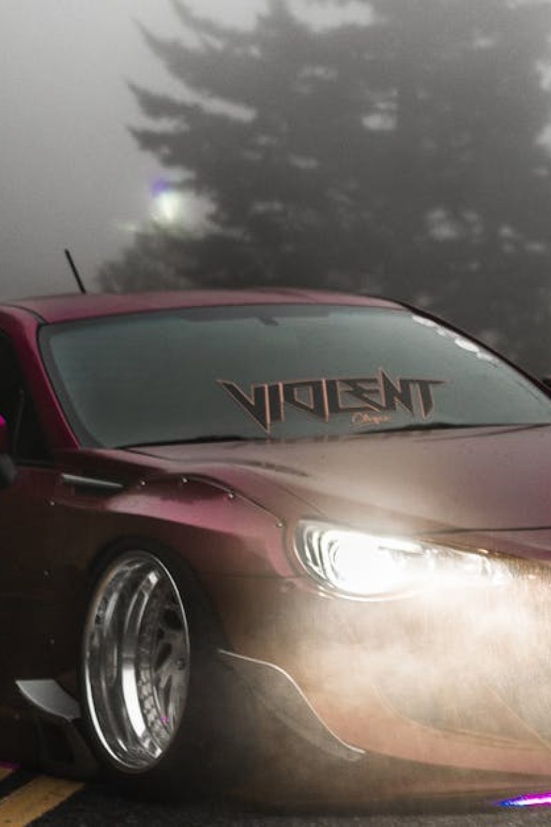 Red sports car with headlights on, driving through a foggy mountain road with pine trees.