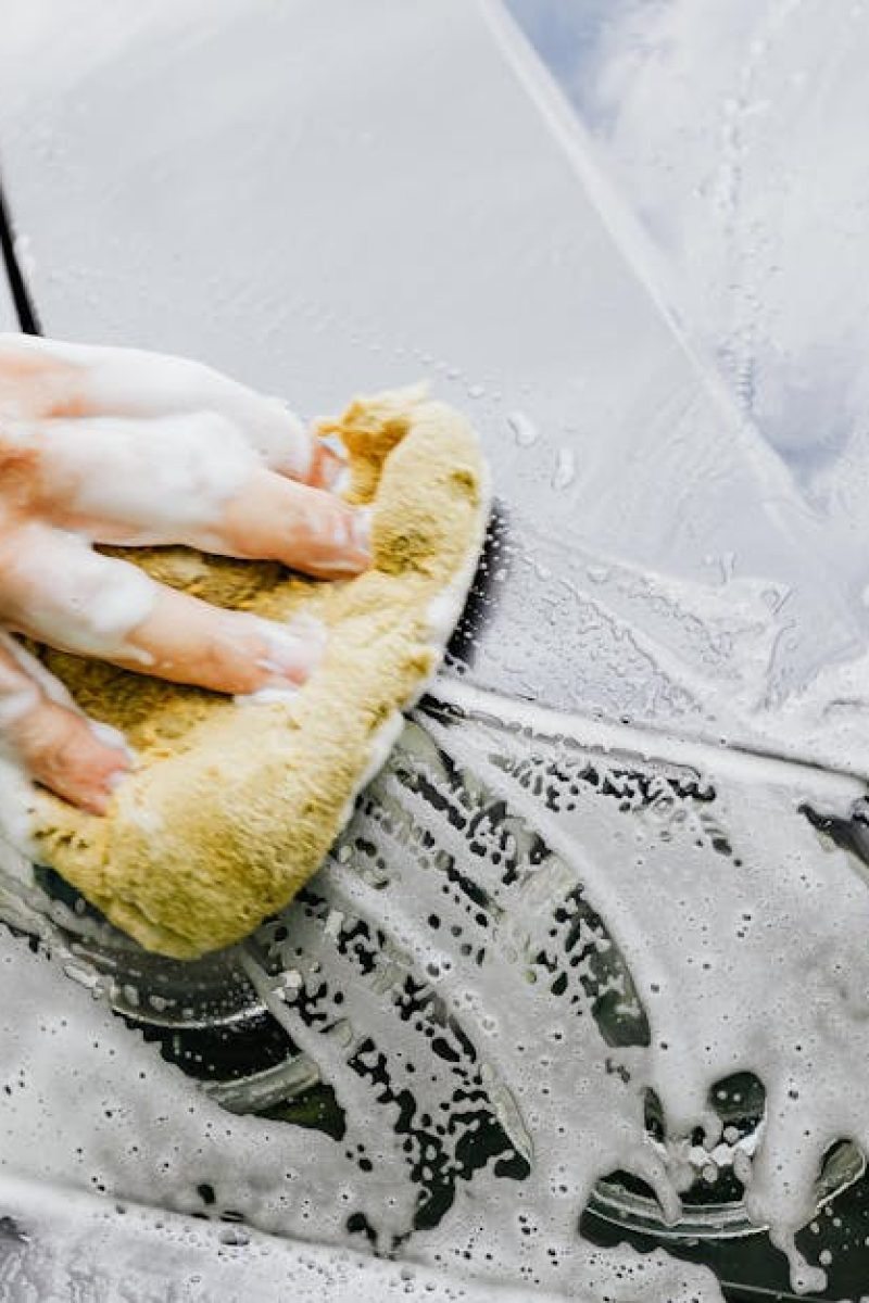 Person washing a black car's headlight with a sponge and soap outdoors.